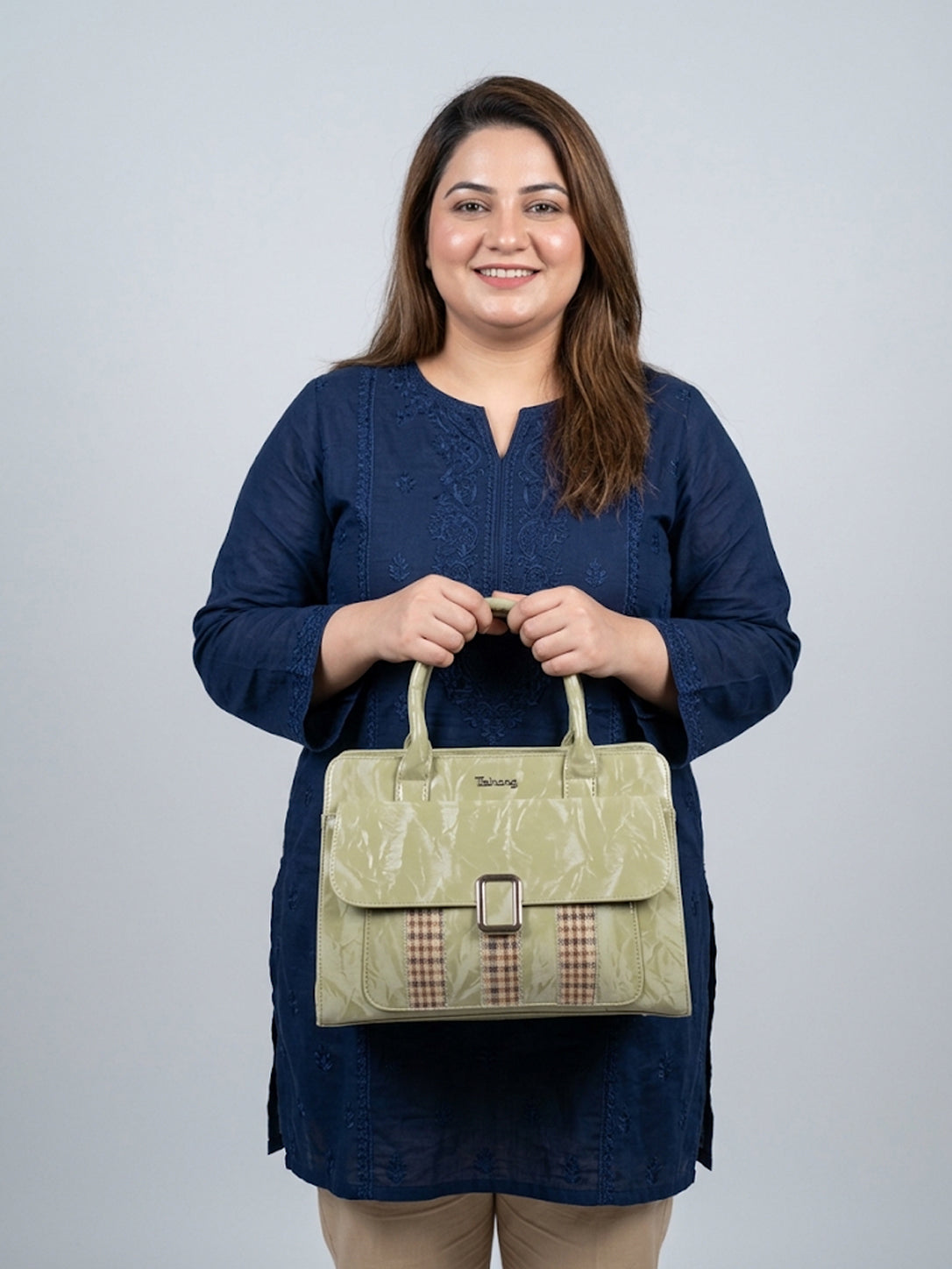 A woman wearing a navy blue tunic holds a light olive green textured handbag with a brown and beige gingham patterned accent strip on the front. The brand name 'Savvy' is visible on the bag against a solid light gray background.